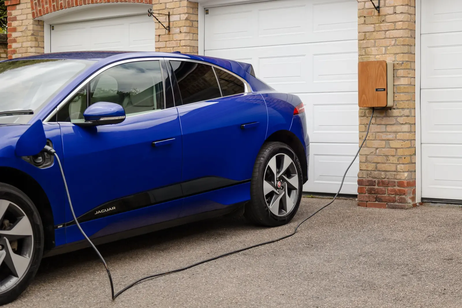A blue electric car plugged into a wall-mounted home charger in front of a garage